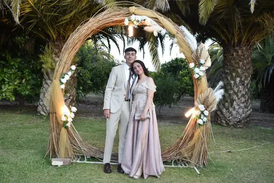 Foto de gala cuarto medio, llegada de alumnos, una pareja tomándose una foto en un arco circular dispuesto, detrás unas plameras